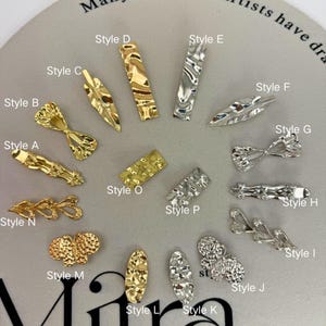 Pode incluir: Uma coleção de encantos de nail art em ouro e prata. Os estilos incluem formas de laço, desenhos de folhas e barras retangulares. Os encantos têm acabamentos texturizados e martelados, adicionando dimensão e interesse visual.