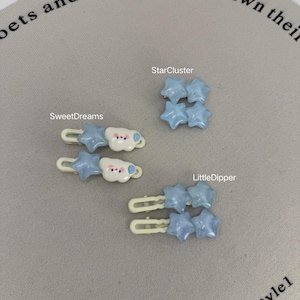 Op de afbeelding: De afbeelding toont haarclips in drie stijlen: "SweetDreams" met een ster- en wolkenontwerp, "StarCluster" met vier blauwe sterren en "LittleDipper" met twee sets blauwe sterren. De clips zijn lichtblauw, wit en crèmekleurig.