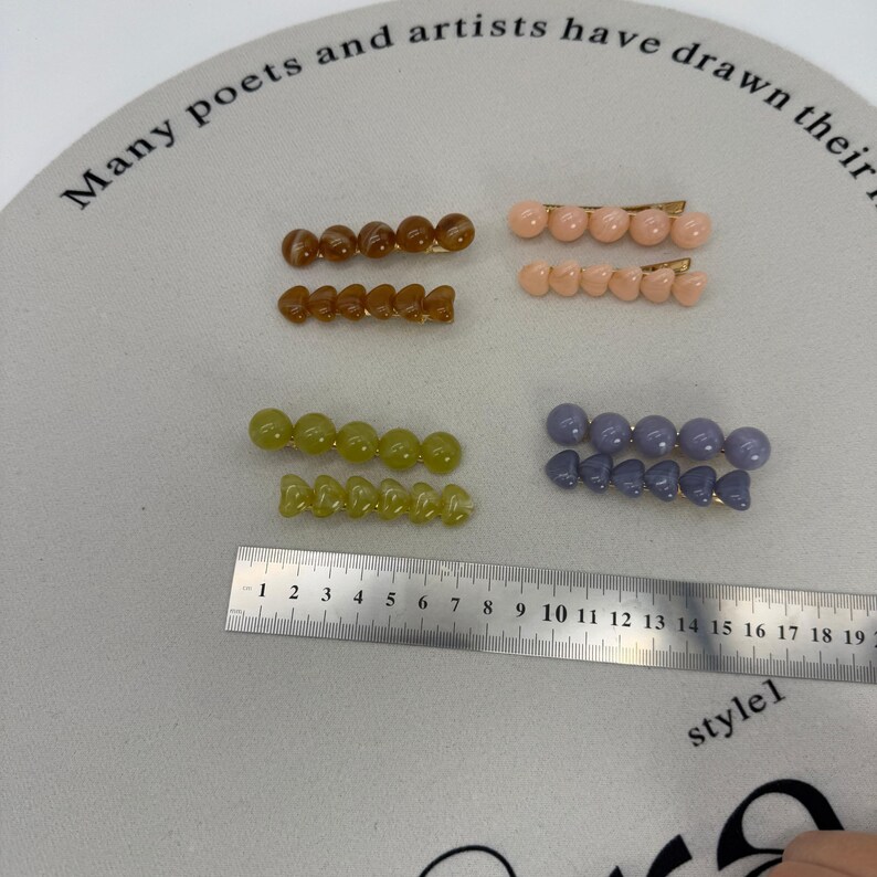 A flat lay photo showing four sets of beaded hair clips arranged on a white background.
Each set consists of two gold-tone alligator clips: one featuring a row of round spherical beads and the other featuring a row of heart-shaped beads.
The sets come in four glossy, jelly-like colors:

Toffee Brown (top left).
Peachy Pink (top right).
Matcha/Olive Green (bottom left).
Dusty Blue/Grey (bottom right).