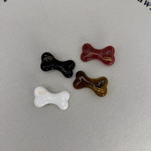 May include: Four bone-shaped decorative items in black, red, brown, and white. Each bone has a glossy finish and the words "Fashion Handmade" printed on it in gold lettering. The items are arranged on a neutral background.