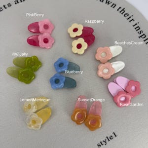 Könnte beinhalten: Eine Sammlung farbenfroher Haarspangen in Blütenform in verschiedenen Farbtönen, darunter Rosa, Grün, Gelb und Orange. Jede Spange hat eine andere Farbkombination, mit Namen wie "PinkBerry" und "KiwiJelly". Das Wort "style1" ist ebenfalls sichtbar.