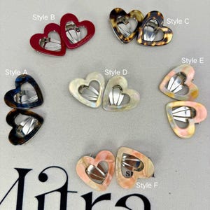 Peut inclure: Une collection de pinces à cheveux en forme de cœur avec des mécanismes à pression en métal, étiquetées de Style A à Style F. Les pinces présentent divers motifs et couleurs, dont le rouge uni, l'écaille, le blanc irisé et des tons pastel marbrés.