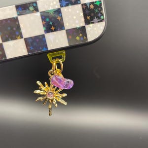 May include: A gold-colored charm with a sunburst design and a purple gemstone, attached to a yellow plastic loop. The charm is connected to a black and white checkered device case with star-shaped accents.