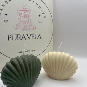 May include: Two seashell-shaped candles, one dark green and one cream-colored, sit in front of a box with the text "Pura Vela" and "Handmade Candles" printed on it. The candles have wicks and are on a white surface.