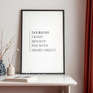 Può includere: Una cornice nera verticale con interno bianco che riporta il testo 2x3 RATIO FRAME MOCKUP PSD WITH SMART OBJECT. Accanto alla cornice si trovano un vaso geometrico blu scuro, un piccolo vaso beige con rami sottili e dei libri bianchi.