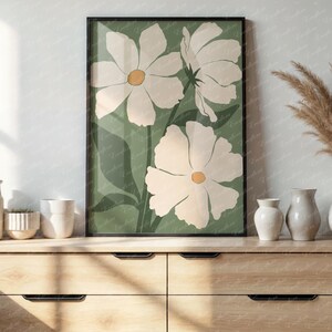 Op de afbeelding: Een ingelijste kunstdruk met drie grote witte bloemen met gele centra op een groene achtergrond. Het kunstwerk wordt tentoongesteld op een houten dressoir, samen met vazen en decoratieve items.