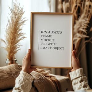 May include: A light wood picture frame containing a white insert with the black text DIN A RATIO FRAME MOCKUP PSD WITH SMART OBJECT. The frame is held upright, showcasing a clean rectangular design suitable for displaying artwork or photography prints.