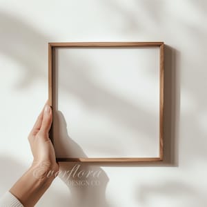Puede incluir: Un marco de fotos cuadrado de madera con un borde fino de madera de color marrón claro natural y un centro blanco vacío. El marco presenta una construcción limpia y sencilla con una textura de veta de madera visible, ofreciendo un soporte neutro.