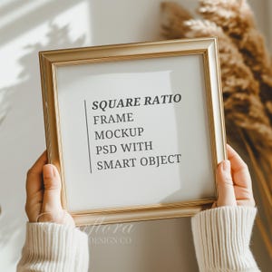 May include: A square gold-coloured picture frame with a white insert featuring the black text: SQUARE RATIO FRAME MOCKUP PSD WITH SMART OBJECT. The frame has a thin, metallic border and is held by hands wearing a beige ribbed knit sweater sleeve.