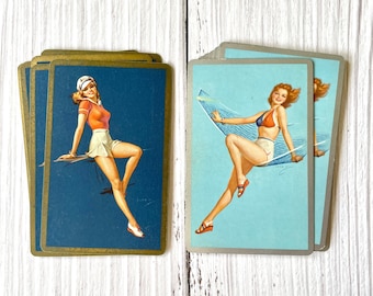 Puede incluir: Dos cartas de juego vintage con ilustraciones de mujeres en traje de baño. La carta de la izquierda tiene un borde dorado y muestra a una mujer con una falda blanca y una blusa roja sentada en un borde. La carta de la derecha tiene un borde plateado y muestra a una mujer con un bikini azul y blanco tumbada en una hamaca.