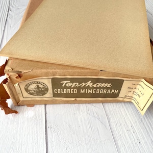 May include: A box of Topsham coloured mimeograph paper with a tan sheet resting on top. The label reads Pejepscot Paper Maine, Topsham Coloured Mimeograph, 500 sheets, size 8 1/2 x 11, 56 Ivory (Tan), and Substance 20. The cardboard box is worn.