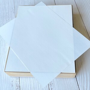 以下が含まれることがあります： 明るい茶色のベースと白い蓋が付いた正方形の箱。半透明の白い紙が箱の上に置かれ、蓋を部分的に覆っています。背景は明るい木目です。