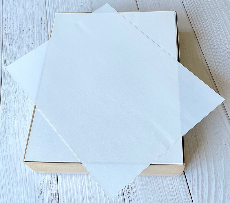 以下が含まれることがあります： 明るい茶色のベースと白い蓋が付いた正方形の箱。半透明の白い紙が箱の上に置かれ、蓋を部分的に覆っています。背景は明るい木目です。