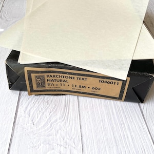 May include: A stack of translucent natural-colored Parchtone text paper sheets resting on a black cardboard box. The box label reads: Parchtone Text Natural, 8 1/2 x 11, 11.8M, 60#, 500 sheets, 1046011, and includes a small logo of a person's face.
