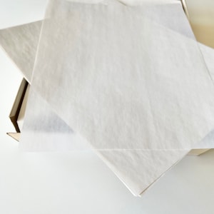 Może przedstawiać: Biały papier jedwabny złożony w kształt kwadratu, w pudełku kartonowym.