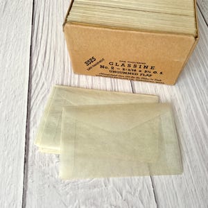 May include: A box containing one thousand ungummed glassine envelopes. The box is brown and has the text "3525 ONE THOUSAND GLASSINE No. 2 - 2-5/16 x 3 1/2 O.S. UNGUMMED FLAP" printed on it. The envelopes are a light beige colour and are stacked neatly inside the box.
