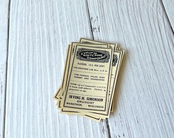 Puede incluir: Un montón de etiquetas de estilo vintage para "Syrup of White Pine Compound". Las etiquetas son de color crema con texto negro y bordes decorativos. El texto incluye información sobre los ingredientes, usos para la tos, resfriados y dolor de garganta, e instrucciones de dosificación.