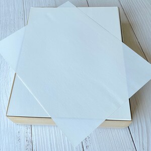 以下が含まれることがあります： 明るい木製のフレームが付いた白い長方形の箱。箱は部分的に開いており、半透明の正方形の紙のシートのスタックが現れています。紙のシートは互いの上に重ねられています。