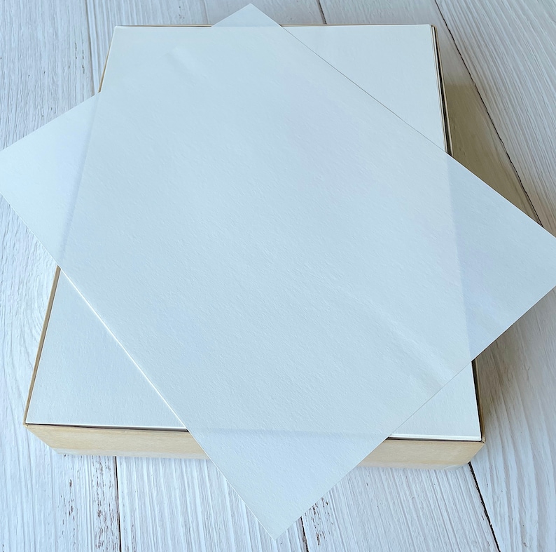 以下が含まれることがあります： 明るい木製のフレームが付いた白い長方形の箱。箱は部分的に開いており、半透明の正方形の紙のシートのスタックが現れています。紙のシートは互いの上に重ねられています。