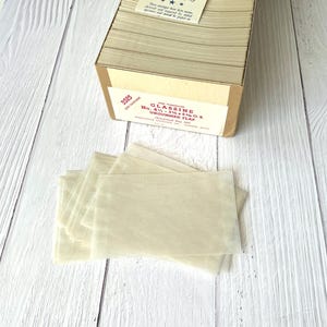 May include: A collection of vintage glassine envelopes, with a box of envelopes in the background. The envelopes are a translucent off-white colour. The box has a label that reads "Glassine No. 4 - 3 1/8 x 5 1/2 O.S. Ungummed Flap."