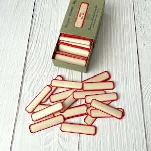 May include: A box of vintage gummed labels with red borders and off-white centers. The box is green with red text and contains a stack of labels. Some labels are scattered on a white wooden surface.