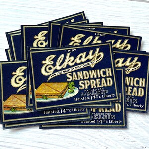 Pode incluir: Uma pilha de etiquetas azuis e douradas para Elkay Sandwich Spread. As etiquetas apresentam um sanduíche com alface e tomate, e o texto "Elkay Sandwich Spread, The Mark of High Quality, Packed for L-Klein, Halsted, 14th & Liberty".