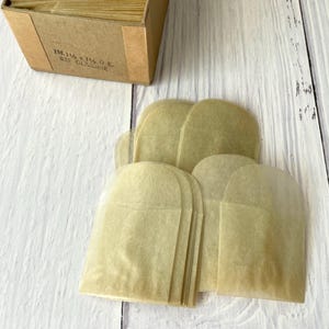 Puede incluir: Una pila de pequeños sobres de papel cristal translúcido de color tostado claro con solapas superiores redondeadas. Una caja de cartón marrón se sitúa sobre los sobres, con el texto impreso 1M-1 1/8 x 1 1/8 O.E. 825 GLASSINE.