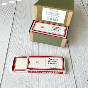 May include: A cardboard carton containing twelve packages of Dennison red bordered gummed labels, item number 31-201-2, with dimensions 3-13/16 inches by 1-9/16 inches. One individual package is removed and placed in front of the open carton.