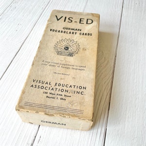 May include: A vintage box of German vocabulary cards from Visual Education Association, Inc. The box is beige with black text and a black and white illustration of an eye with books and a sunburst.