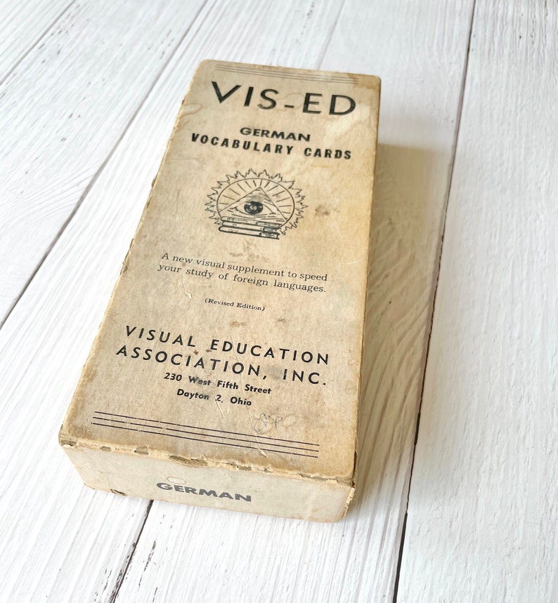 May include: A vintage box of German vocabulary cards from Visual Education Association, Inc. The box is beige with black text and a black and white illustration of an eye with books and a sunburst.