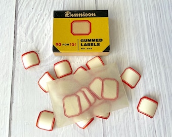 May include: A vintage box of Dennison gummed labels, with the text "90 for 15¢". The box is yellow and black, with a red-bordered label design. Loose labels with red borders and cream centers are scattered around.