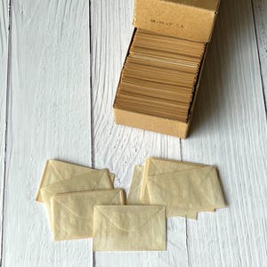 May include: A cardboard box filled with a stack of small, translucent, light tan paper envelopes. The box lid is open and features the printed text 1M - 1 5/8 x 2 1/2 O.S. Several additional empty envelopes are arranged in a cluster below the box.