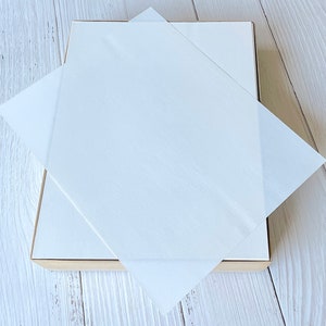 以下が含まれることがあります： 明るい木製のフレームが付いた白い長方形の箱。半透明の白い紙が対角線上に置かれ、一部を覆っています。背景は明るい色の木製の表面です。