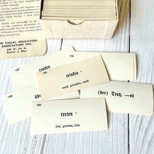 May include: Close-up of a set of flashcards with German vocabulary words. The cards are white with black text. The words on the cards include "troden", "trinken", "treten", and "(der) Tros".