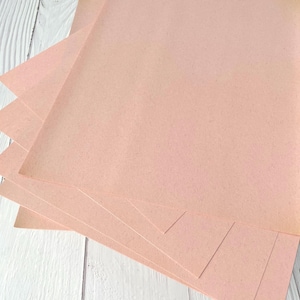 May include: A stack of light pink paper sheets, possibly cardstock, arranged on a white wooden surface. The paper has a slightly textured appearance and a warm, inviting color. The sheets are neatly stacked, with some edges visible.