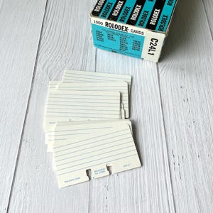 May include: A box of 1000 Rolodex cards with a teal and white label featuring the text Rolodex and C24L1. A stack of white index cards with blue horizontal lines and a notched bottom edge sits in front of the box, displaying the Rolodex Corp branding.