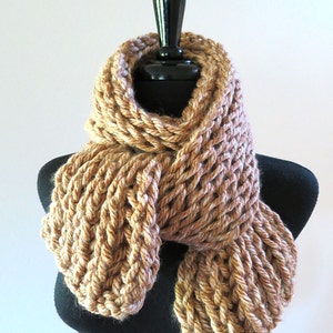 May include: A chunky, hand-knitted scarf in shades of tan and beige, displayed on a black mannequin. The scarf features a thick, open weave design, creating a textured and cozy appearance. The scarf is designed to be worn around the neck.