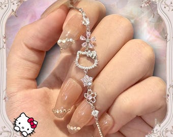 May include: A silver charm bracelet with a Hello Kitty charm, pink flower charms, and clear rhinestones. The bracelet is on a hand with long, light brown, square-shaped nails with glitter accents.