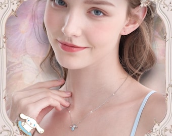 May include: A woman wearing a light blue tank top with a silver necklace featuring a small blue and white charm. The charm is a cartoon character with a blue bow and a white body. The woman is wearing a silver ring with a cartoon character on it.