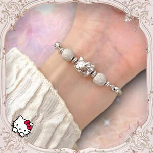 May include: A silver charm bracelet with a Hello Kitty charm and silver beads. The bracelet is on a person's wrist.