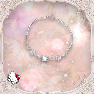May include: A silver bracelet with a pink Hello Kitty charm and pink flower accents. The bracelet is on a pink and white background.