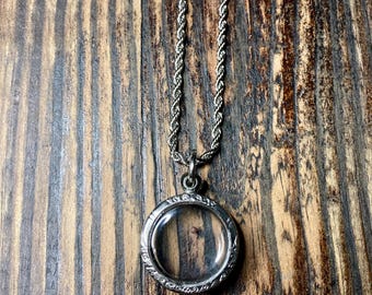 May include: Silver locket necklace with a clear glass centre, suspended from a matching rope chain. The locket has a decorative, patterned border. The necklace is displayed against a rustic, wooden background.