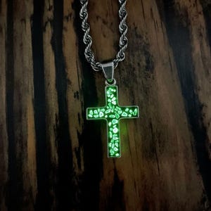 May include: A silver-toned necklace with a cross pendant. The cross is filled with glowing green stones and is suspended from a rope chain. The background is a dark wood grain.