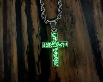 May include: A silver necklace featuring a cross pendant filled with glowing green stones. The cross hangs from a rope chain. The background is a dark wood grain pattern.
