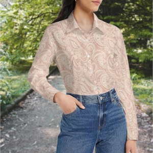 Pode incluir: Camisa de botão de manga comprida com estampa paisley em tons de bege e rosa. Combinada com jeans azuis. O fundo apresenta uma cena externa desfocada com um caminho e árvores.