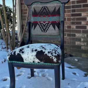 May include: A black wooden chair with a carved back and a cowhide seat. The chair is upholstered with a patterned fabric featuring geometric designs in shades of brown, gray, red, and blue.