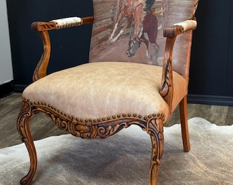 May include: A vintage armchair with a western-themed design. The chair features a light brown leather seat and a backrest with a painting of a cowboy riding a bucking bronco. The wooden frame has ornate carvings and armrests.