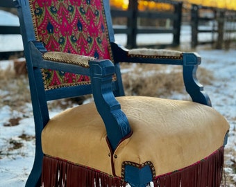 May include: A vintage chair with a dark blue frame, a pink and gold patterned fabric back, and a tan leather seat with brown fringe.