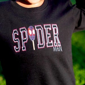 May include: Black long-sleeved shirt with the word "SPYDER" in white, outlined in black and pink, with a Spiderman mask graphic. The word "MAN" is below the word "DER".