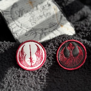 May include: Two embroidered patches on a black fabric background. The patch on the left is red and white with a stylized sword and wings. The patch on the right is red and black with a stylized rebel alliance symbol.